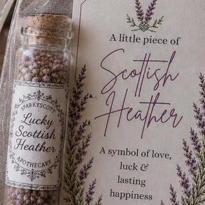 May include: A clear glass vial with a cork stopper filled with small purple heather flowers, accompanied by a rectangular card featuring purple heather illustrations and the text: A little piece of Scottish Heather, A symbol of love, luck & lasting happiness.