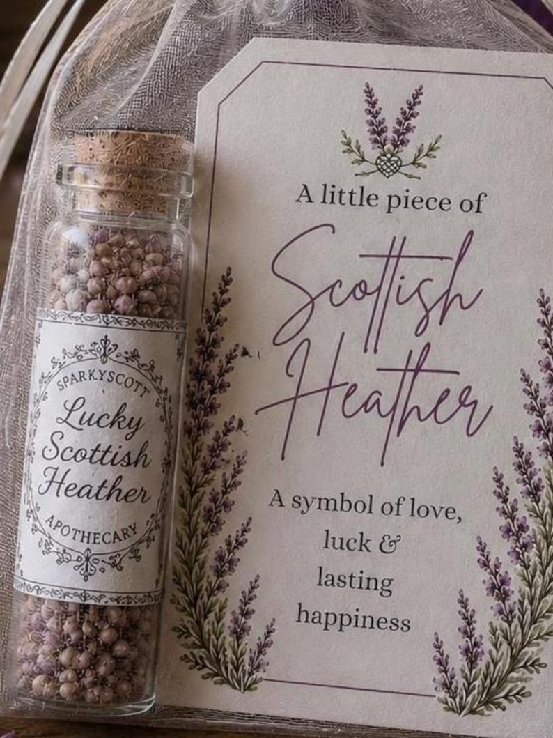 May include: A clear glass vial with a cork stopper filled with small purple heather flowers, accompanied by a rectangular card featuring purple heather illustrations and the text: A little piece of Scottish Heather, A symbol of love, luck & lasting happiness.