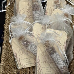 May include: Several small glass vials filled with dried purple heather, each sealed with a cork stopper and tucked into a white organza drawstring bag. Each bag includes a beige paper tag printed with the text A little piece of Scottish Heather.