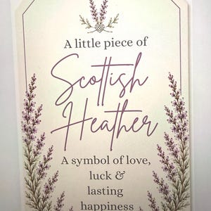 May include: A rectangular off-white sign featuring purple heather illustrations on the sides and top. The text reads: A little piece of Scottish Heather, A symbol of love, luck & lasting happiness. The sign has a thin purple border and rounded top corners.