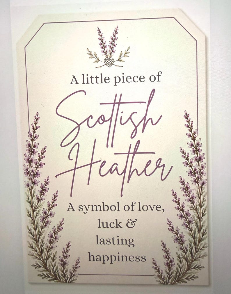 May include: A rectangular off-white sign featuring purple heather illustrations on the sides and top. The text reads: A little piece of Scottish Heather, A symbol of love, luck & lasting happiness. The sign has a thin purple border and rounded top corners.