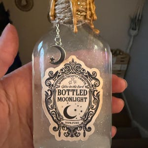 May include: A clear glass bottle filled with glittery liquid, featuring a label that reads Sparklyscotty Crafts Glow in the dark BOTTLED MOONLIGHT 100% PURE. The neck is wrapped in twine with gold wax drips and a silver crescent moon and star charm.