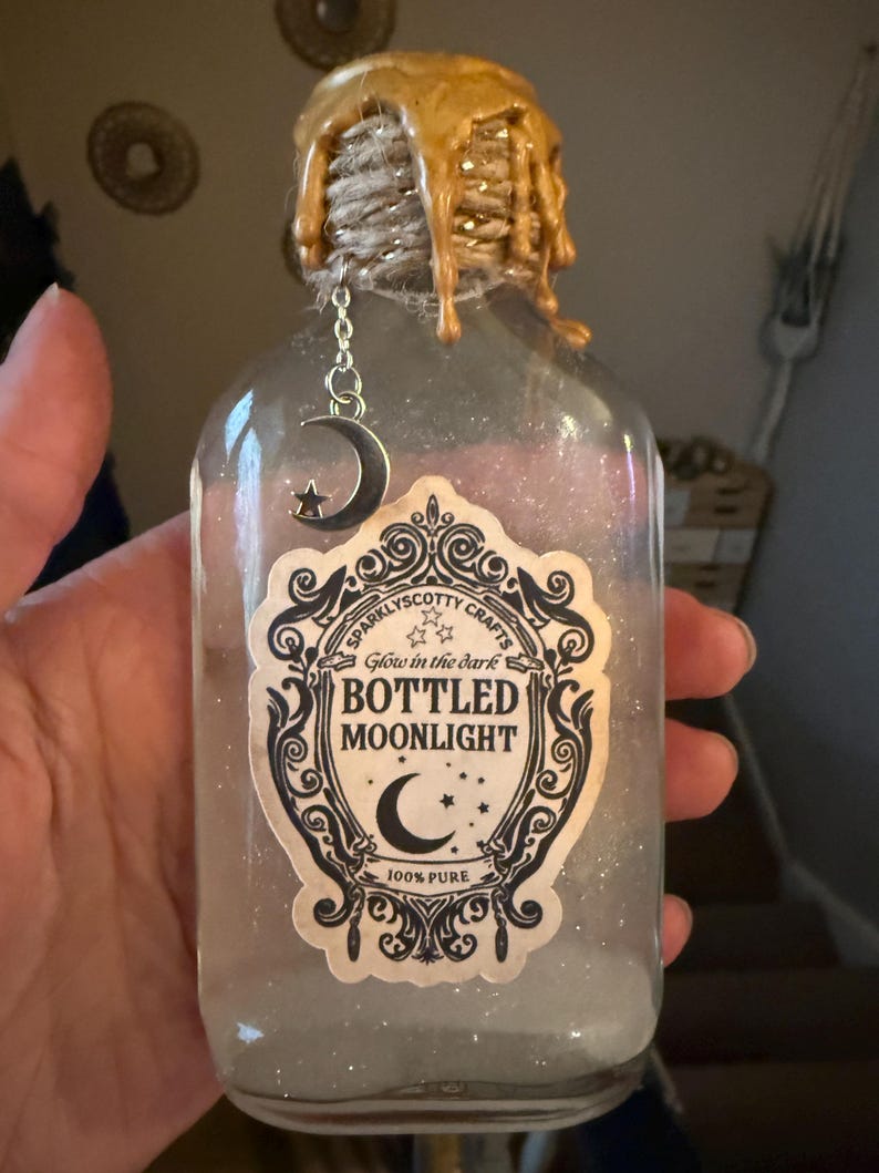 May include: A clear glass bottle filled with glittery liquid, featuring a label that reads Sparklyscotty Crafts Glow in the dark BOTTLED MOONLIGHT 100% PURE. The neck is wrapped in twine with gold wax drips and a silver crescent moon and star charm.