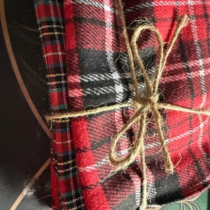 K&ouml;nnte beinhalten: Ein Stapel rot-schwarz karierter Stoffquadrate, mit Bindfaden zusammengebunden. Das Karomuster enth&auml;lt wei&szlig;e Akzente. Der Stoff ist gefaltet und geb&uuml;ndelt, was auf ein Set von Untersetzern oder kleinen Dekorationsartikeln hindeutet.