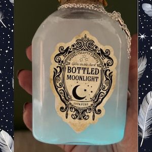 Könnte beinhalten: Eine klare Glasflasche gefüllt mit einer hellblauen Flüssigkeit, beschriftet mit "Bottled Moonlight". Das Etikett ist mit einem Mond und Sternen verziert. Eine silberne Kette ist am Korken befestigt. Die Flasche leuchtet von unten.