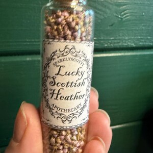 May include: A clear glass vial with a cork stopper filled with small, dried purple and green heather flowers. A rectangular paper label is attached to the front, featuring an ornate black border and the text Lucky Scottish Heather Apothecary.
