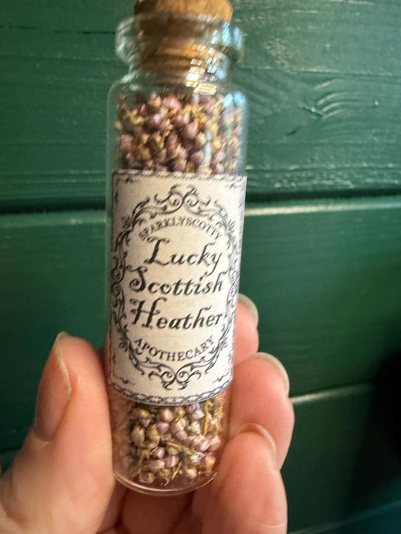 May include: A clear glass vial with a cork stopper filled with small, dried purple and green heather flowers. A rectangular paper label is attached to the front, featuring an ornate black border and the text Lucky Scottish Heather Apothecary.