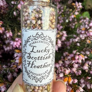 May include: A clear glass vial filled with dried light purple heather flowers, sealed with a cork stopper. A white rectangular label features black decorative borders and the text: Sparklyscotty Lucky Scottish Heather Apothecary.