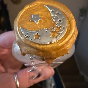 May include: A bottle stopper featuring a circular gold wax seal top with a silver-coloured crescent moon and star motif embossed on the surface. The gold wax has a textured, dripped appearance around the edges, and a small silver moon charm hangs.