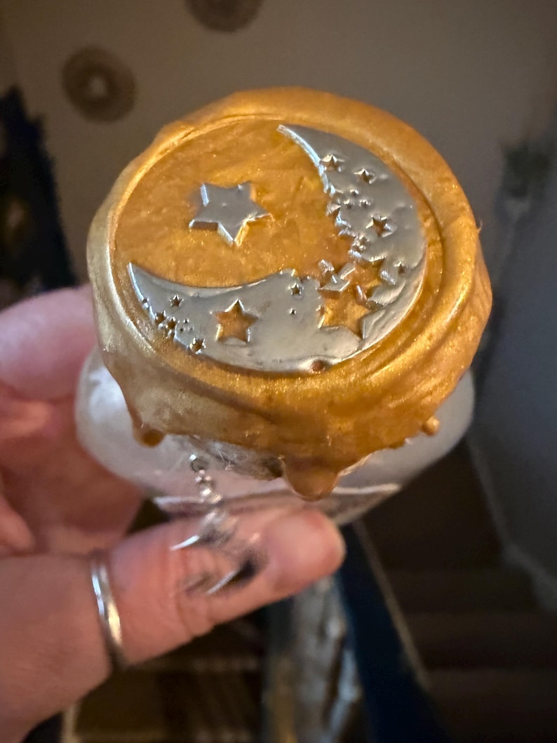 May include: A bottle stopper featuring a circular gold wax seal top with a silver-coloured crescent moon and star motif embossed on the surface. The gold wax has a textured, dripped appearance around the edges, and a small silver moon charm hangs.