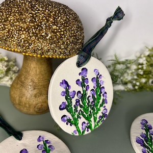 May include: Three round white ceramic ornaments with purple and green painted heather flowers. Each ornament has a green and black plaid ribbon attached.