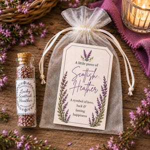 May include: A clear glass vial with a cork stopper filled with small purple Scottish heather flowers, accompanied by a sheer white organza drawstring pouch featuring a tag that reads A little piece of Scottish Heather, A symbol of love, luck & lasting happiness.