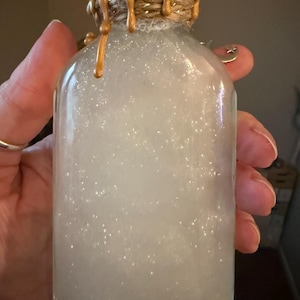 May include: A clear glass bottle filled with a white, shimmering, pearlescent liquid. The neck of the bottle is wrapped in natural jute twine, topped with metallic gold wax drips that cascade down the sides. A small silver crescent moon charm is attached.