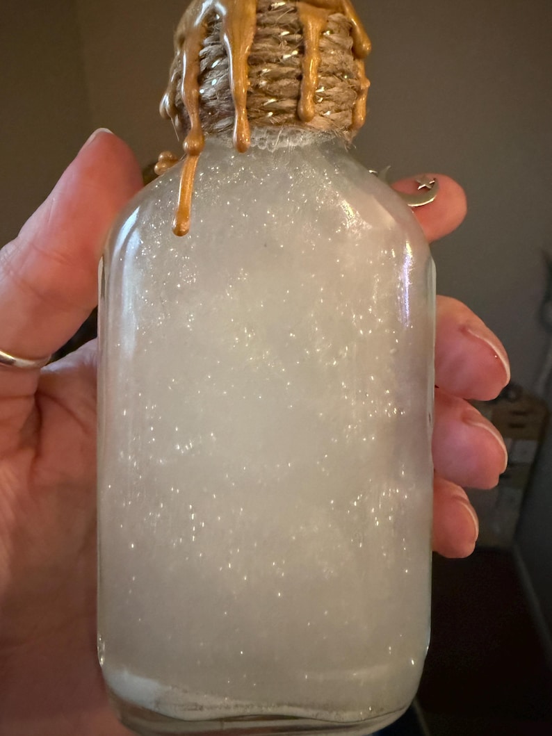 May include: A clear glass bottle filled with a white, shimmering, pearlescent liquid. The neck of the bottle is wrapped in natural jute twine, topped with metallic gold wax drips that cascade down the sides. A small silver crescent moon charm is attached.
