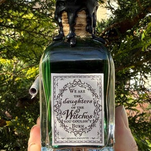 Puede incluir: Una botella de vidrio verde con un sello de cera negra y un tapón de corcho. La botella tiene una etiqueta que dice "We are the daughters of the Witches you couldn't burn". La botella se sostiene contra un fondo de follaje verde.
