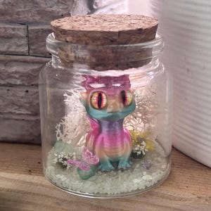 Op de afbeelding: Een heldere glazen pot met een kurken deksel, met een kleurrijk, 3D-geprint drakenfiguur. De draak is voornamelijk roze en blauw, met gele ogen. De pot bevat ook wit mos, kleine bloemen en een roze vlinderfiguur, waardoor een miniatuur terrarium ontstaat.