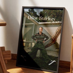 May include: Framed print with the text "Drew Starkey." The image depicts a person in a patterned sweater, green trousers, and black and white trainers. The background includes a house and a tree.