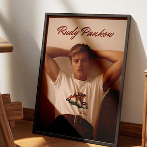 May include: Framed poster featuring a person in a white t-shirt with a graphic design and a brown belt. The poster has the name "Rudy Pankow" in a cursive font at the top. The frame is black, and the background is a light beige color.
