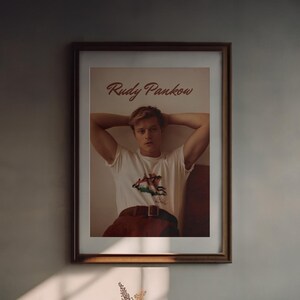 May include: A framed print featuring the name "Rudy Pankow" in a cursive font. The print showcases a person wearing a white t-shirt with a graphic design, paired with red pants and a brown belt. The image is in a brown wooden frame.