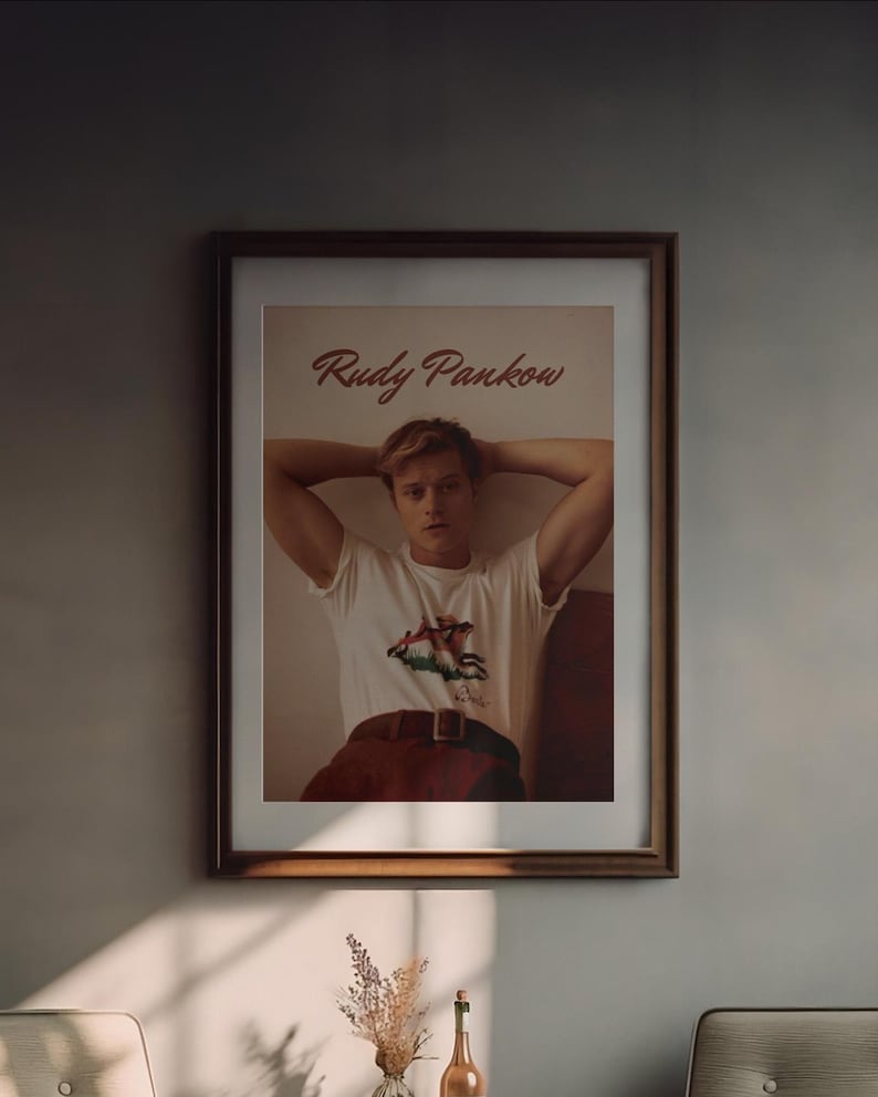 May include: A framed print featuring the name "Rudy Pankow" in a cursive font. The print showcases a person wearing a white t-shirt with a graphic design, paired with red pants and a brown belt. The image is in a brown wooden frame.