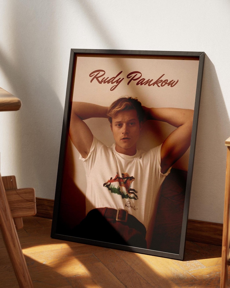 May include: Framed poster featuring a person in a white t-shirt with a graphic design and a brown belt. The poster has the name "Rudy Pankow" in a cursive font at the top. The frame is black, and the background is a light beige color.