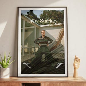 May include: A framed poster with the name 'Drew Starkey' at the top. The subject wears a patterned sweater, green trousers, and black and white trainers. The poster is on a wooden cabinet with a plant and a wooden hand.
