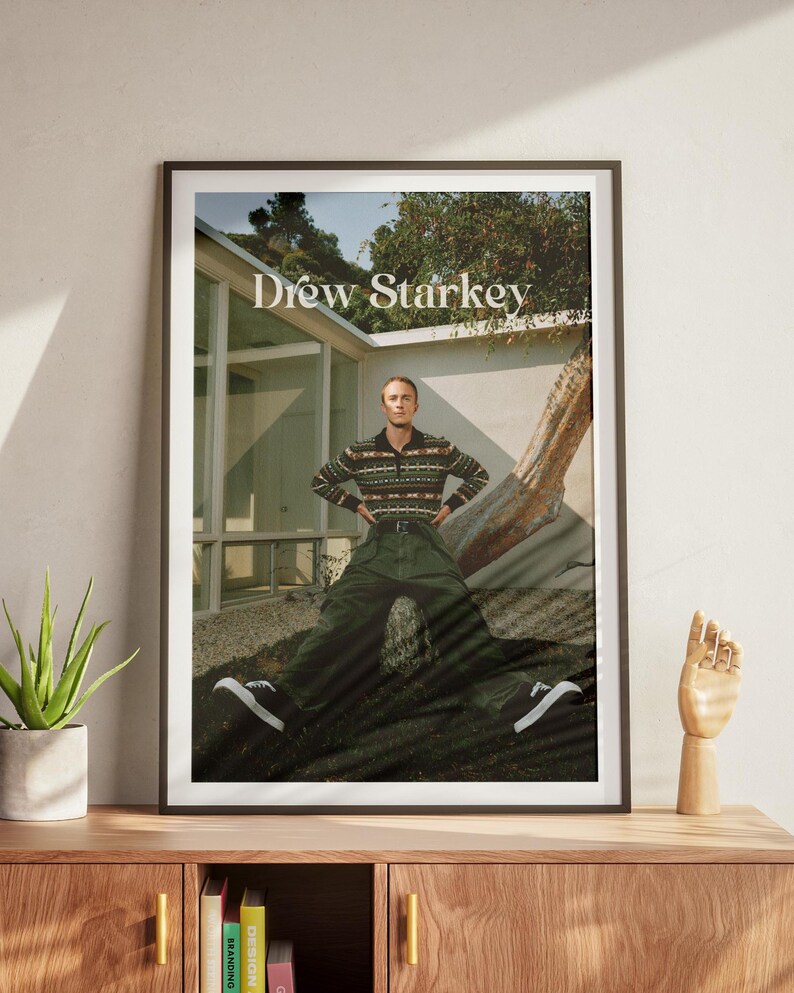 May include: A framed poster with the name 'Drew Starkey' at the top. The subject wears a patterned sweater, green trousers, and black and white trainers. The poster is on a wooden cabinet with a plant and a wooden hand.