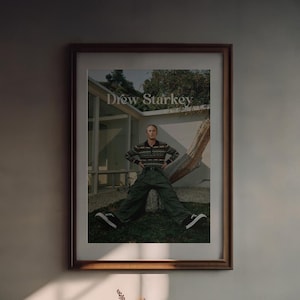 May include: Framed print featuring Drew Starkey. The image shows Starkey seated against a tree, wearing a patterned sweater, green trousers, and black trainers. The print is in a brown wooden frame, displayed on a neutral-coloured wall.
