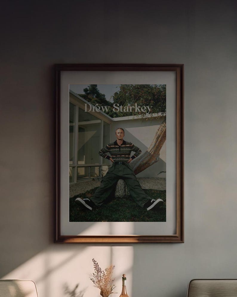 May include: Framed print featuring Drew Starkey. The image shows Starkey seated against a tree, wearing a patterned sweater, green trousers, and black trainers. The print is in a brown wooden frame, displayed on a neutral-coloured wall.