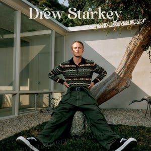 May include: A person wearing a patterned sweater, dark green trousers, and black trainers. The text "Drew Starkey" is displayed above. The person is sitting on a rock in a grassy area with a tree and a modern building in the background.