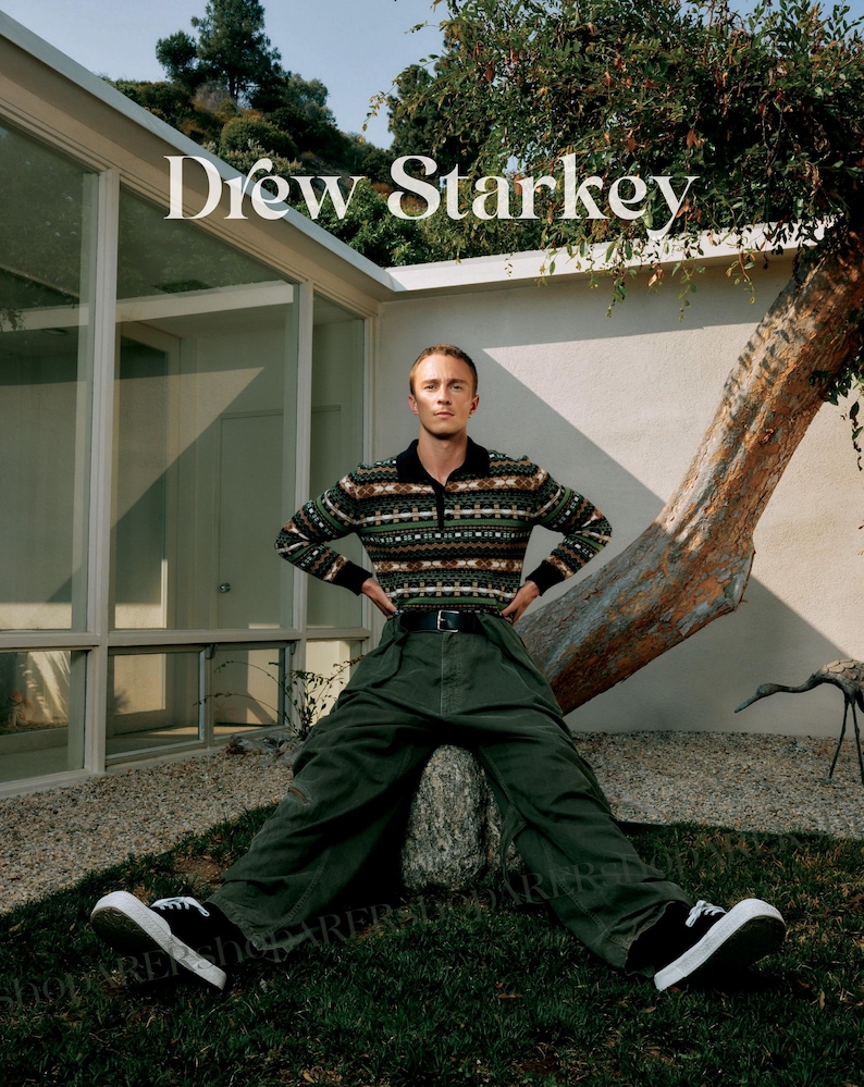 May include: A person wearing a patterned sweater, dark green trousers, and black trainers. The text "Drew Starkey" is displayed above. The person is sitting on a rock in a grassy area with a tree and a modern building in the background.