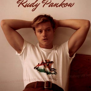 May include: A white t-shirt with a graphic of a fox and a house, paired with brown pants and a brown belt. The name "Rudy Pankow" is written in brown script above the person.