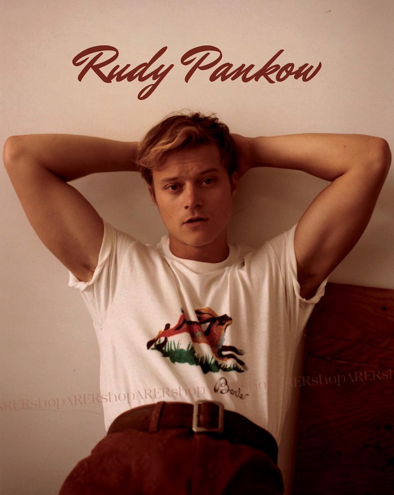 May include: A white t-shirt with a graphic of a fox and a house, paired with brown pants and a brown belt. The name "Rudy Pankow" is written in brown script above the person.