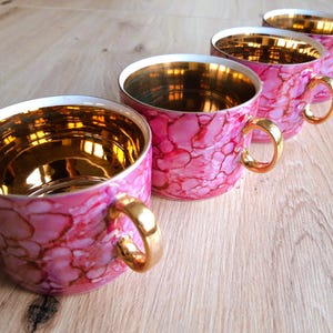 May include: Four pink teacups with gold rims and handles are arranged on a light wood surface. The cups have a marbled design in shades of pink and red. The interior of each cup is gold.