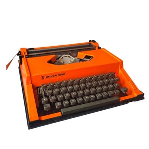 Puede incluir: Máquina de escribir Rover 1000 naranja. El teclado negro presenta una disposición QWERTY. El cuerpo naranja brillante lleva la inscripción «Rover 1000». El diseño es retro y funcional, ideal para escribir texto.
