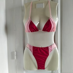 May include: A two-piece bikini set on a mannequin. The top is a triangle style with halter straps, and the bottoms are high-cut. The colour scheme includes shades of pink and red, with a striped design.