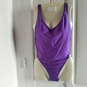 May include: A purple one-piece swimsuit with a scoop neck and thin straps. The swimsuit is on a white mannequin.