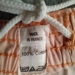 Puede incluir: Primer plano de una tela de color melocot&oacute;n con un cord&oacute;n blanco y una etiqueta blanca que dice "Made in France 100% Coton". La etiqueta tambi&eacute;n tiene una etiqueta de cuidado con un s&iacute;mbolo de 60 grados, un tri&aacute;ngulo y un c&iacute;rculo con una cruz dentro.