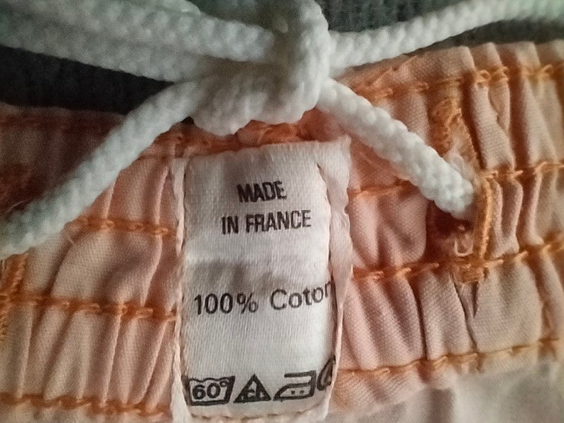 Puede incluir: Primer plano de una tela de color melocot&oacute;n con un cord&oacute;n blanco y una etiqueta blanca que dice "Made in France 100% Coton". La etiqueta tambi&eacute;n tiene una etiqueta de cuidado con un s&iacute;mbolo de 60 grados, un tri&aacute;ngulo y un c&iacute;rculo con una cruz dentro.