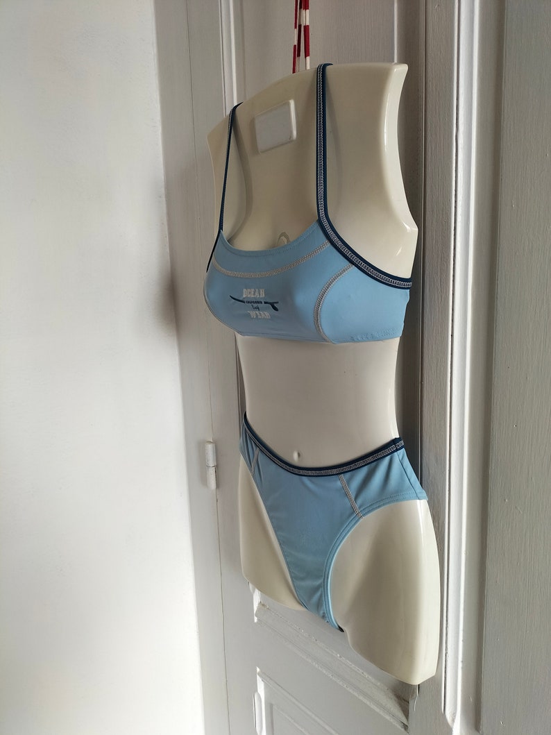 Op de afbeelding: Lichtblauwe bikiniset op een mannequin. De top heeft dunne bandjes en de tekst "OCEAN" met een surfplankafbeelding. De randen zijn afgewerkt met marineblauwe en lichtbeige stiksels. De bijpassende broek is ook lichtblauw.