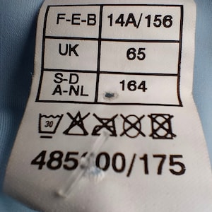 Op de afbeelding: Een wit kledinglabel met zwarte tekst en symbolen. Het label bevat maat- en onderhoudsinstructies. Het label heeft de volgende tekst: F-E-B 14A/156, UK 65, S-D A-NL 164, 48500/175 en wasvoorschriften.