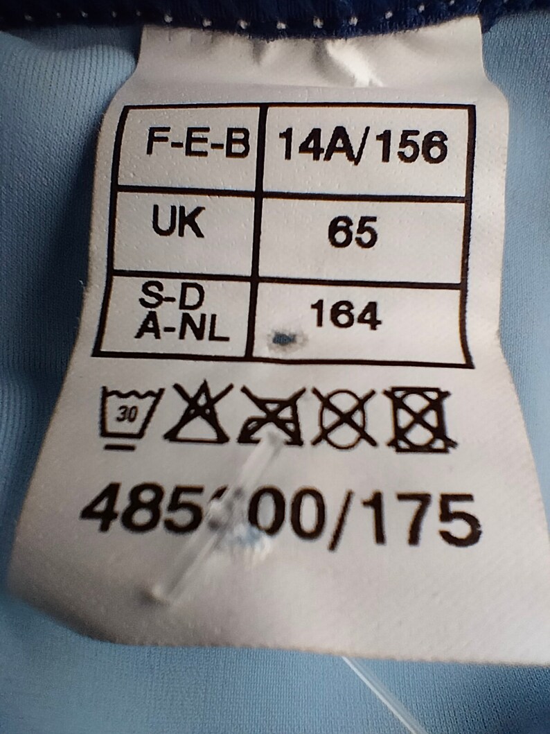 Op de afbeelding: Een wit kledinglabel met zwarte tekst en symbolen. Het label bevat maat- en onderhoudsinstructies. Het label heeft de volgende tekst: F-E-B 14A/156, UK 65, S-D A-NL 164, 48500/175 en wasvoorschriften.