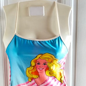 Puede incluir: Un traje de ba&ntilde;o azul y rosa con ribete blanco y un gr&aacute;fico de una Barbie rubia con una camisa a rayas rosa y blanca.