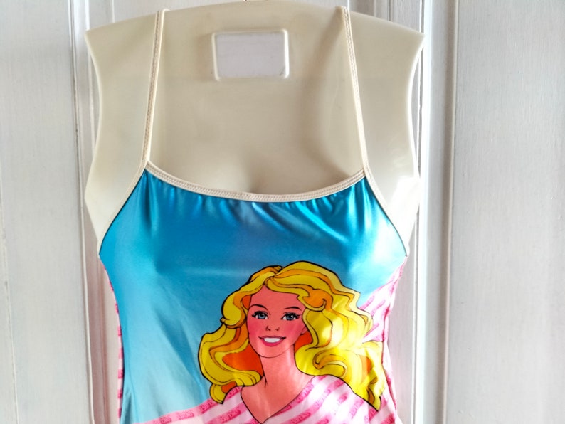 Puede incluir: Un traje de ba&ntilde;o azul y rosa con ribete blanco y un gr&aacute;fico de una Barbie rubia con una camisa a rayas rosa y blanca.