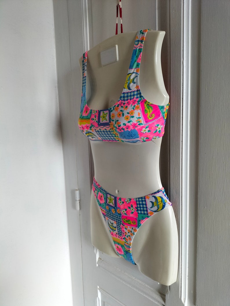 Puede incluir: Un traje de ba&ntilde;o de dos piezas con un dise&ntilde;o de patchwork vibrante. La parte superior e inferior presentan una mezcla de estampados florales, cuadros y geom&eacute;tricos en rosa, azul, amarillo y verde. El traje de ba&ntilde;o se exhibe en un maniqu&iacute; blanco.