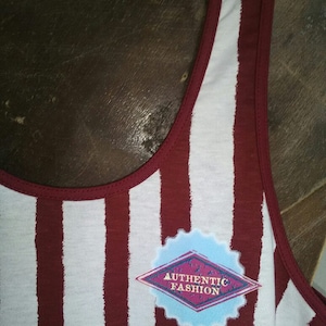 May include: A white and maroon striped tank top with a diamond-shaped patch that says "AUTHENTIC FASHION" in gold lettering.