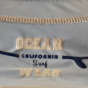 Op de afbeelding: Lichtblauw shirt met de tekst "OCEAN CALIFORNIA Surf WEAR" in witte blokletters. Een donkerblauwe surfboard-afbeelding staat links en rechts van de tekst. Het shirt heeft een lichtbeige kraag.