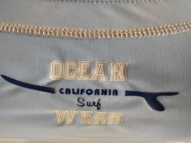 Op de afbeelding: Lichtblauw shirt met de tekst "OCEAN CALIFORNIA Surf WEAR" in witte blokletters. Een donkerblauwe surfboard-afbeelding staat links en rechts van de tekst. Het shirt heeft een lichtbeige kraag.