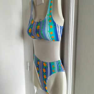 Op de afbeelding: Een blauw-wit gestreept badpak met oranje bloemen. Het badpak heeft een haltertop en een hoog uitgesneden onderkant.