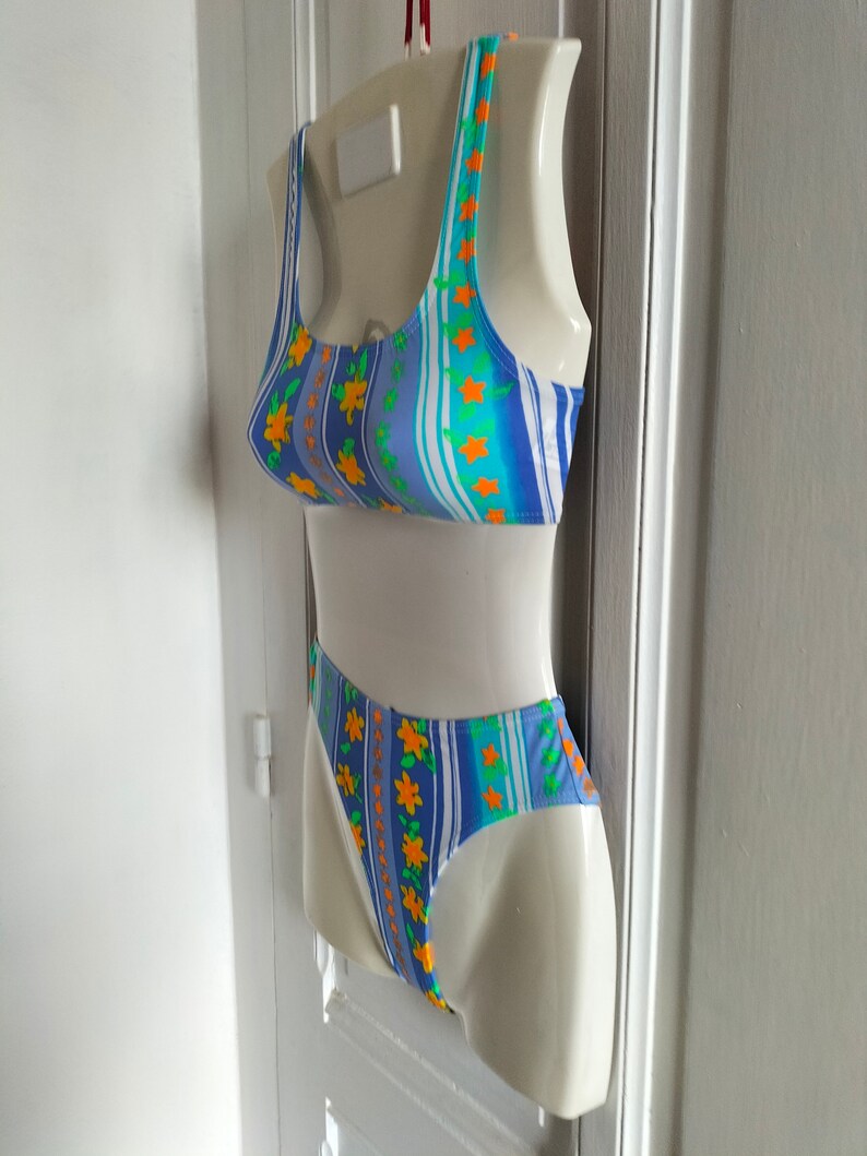 Op de afbeelding: Een blauw-wit gestreept badpak met oranje bloemen. Het badpak heeft een haltertop en een hoog uitgesneden onderkant.